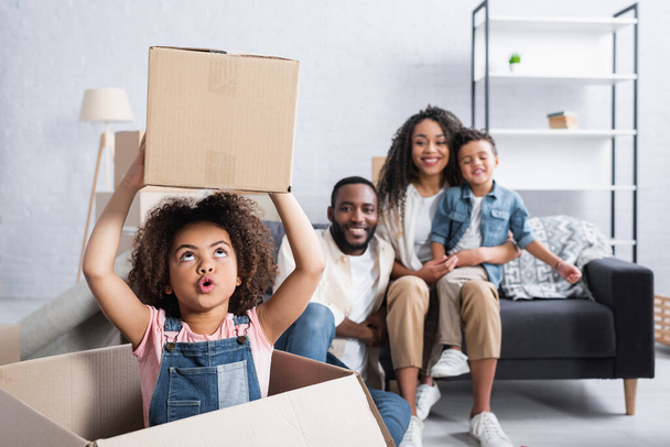 excited african american child holding cardboard box near blurred family - Photo, Image