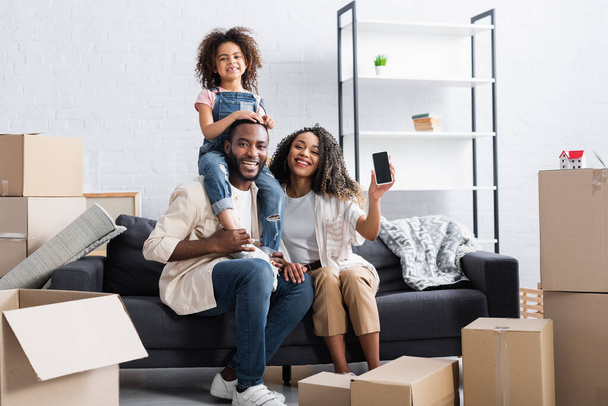 fröhliche afrikanisch-amerikanische Frau zeigt Handy mit leerem Bildschirm nahe Familie in neuer Wohnung  - Foto, Bild