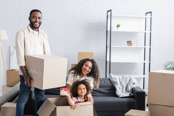 happy african american girl in cardboard box holding house model near parents in new apartment - Photo, Image