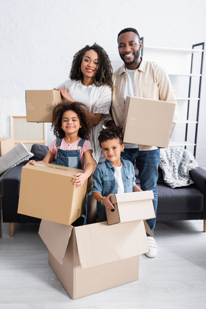 familia afroamericana feliz con cajas de cartón mirando a la cámara en nuevo apartamento  - Foto, Imagen