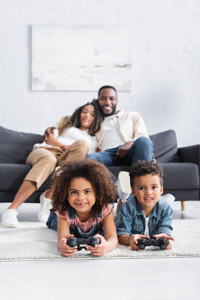 KYIV, UKRAINE - JANUARY 25, 2021: african american brother and sister gaming on floor near blurred parents - Foto, Imagen