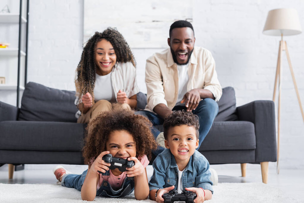 KYIV, UCRANIA - 25 de enero de 2021: padres afroamericanos emocionados cerca de niños jugando videojuegos en el suelo - Foto, imagen