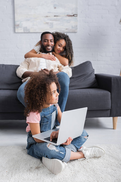 happy african american woman embracing husband near daughter sitting on floor with laptop - Foto, imagen
