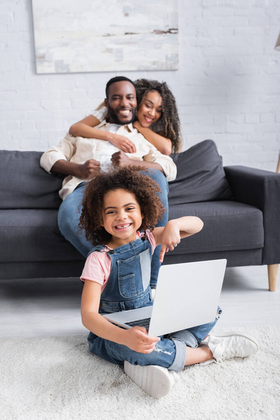 happy african american girl pointing at laptop while sitting on floor near blurred parents - Foto, Bild