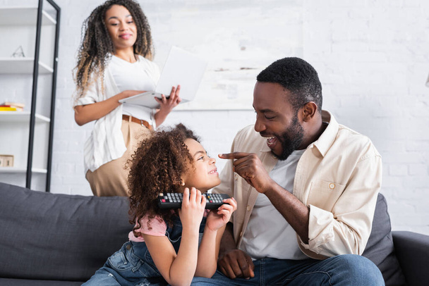 cheerful african american man touching nose of daughter near blurred wife with laptop - Photo, Image