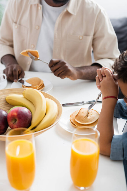 abgeschnittene Ansicht eines afrikanisch-amerikanischen Mannes mit Sohn in der Nähe von Pfannkuchen, Früchten und Orangensaft - Foto, Bild