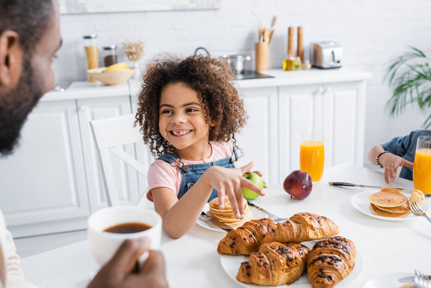 cheerful african american girl pointing at croissants during breakfast with brother and blurred dad - Photo, Image