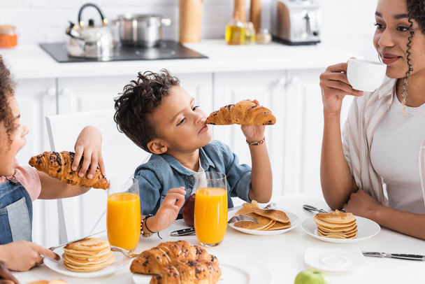 african american kids eating croissants near happy mom drinking coffee during breakfast - Fotoğraf, Görsel