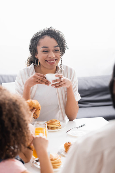 smiling african american woman holding cup of coffee during breakfast with family - Foto, Imagem