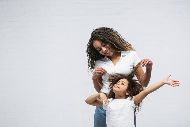 radostná žena dotýkající se vlasů kudrnaté africké americké dcery na šedé - Fotografie, Obrázek