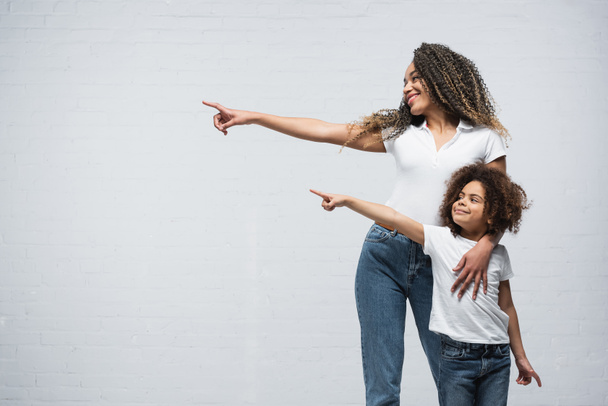 sorridente afro-americano mãe e filha olhando para o lado e apontando com os dedos no cinza - Foto, Imagem
