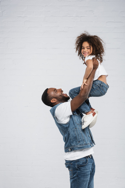 joyful african american man raising happy child on grey - Foto, Bild