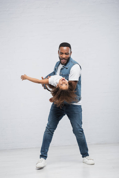 cheerful african american man holding excited daughter in hands on grey - Photo, Image