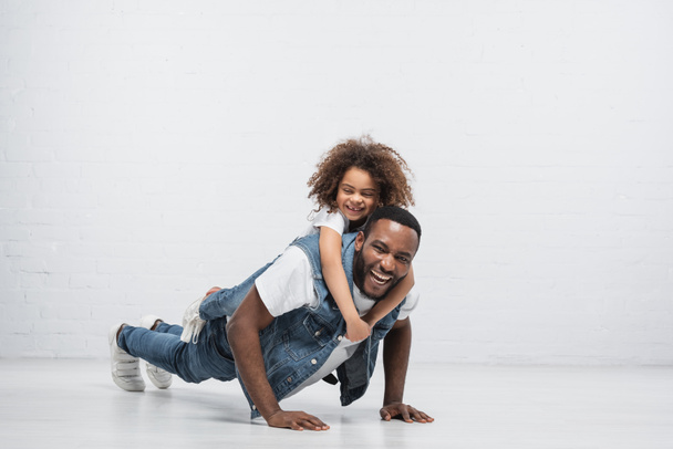 allegro afro americano uomo fare flessioni con felice figlia cavalluccio lui su grigio - Foto, immagini