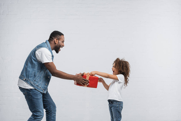 excited african american man giving red gift box to amazed daughter on grey - Foto, Bild