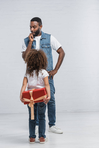 back view of african american kid holding red gift box behind back near thoughtful dad on grey - 写真・画像