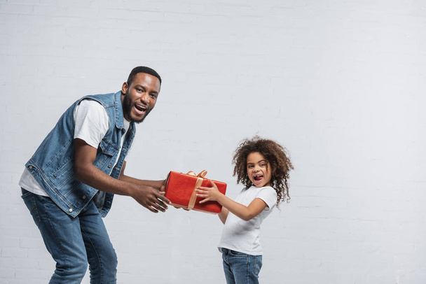 έκπληκτος Αφροαμερικάνος πατέρας και κόρη κρατώντας κόκκινο κουτί δώρου σε γκρι - Φωτογραφία, εικόνα
