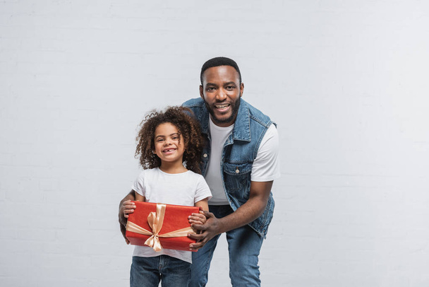 ευχαριστημένος αφρικανός Αμερικανός παιδί δείχνει κόκκινο κουτί δώρου κοντά χαρούμενος μπαμπάς σε γκρι - Φωτογραφία, εικόνα