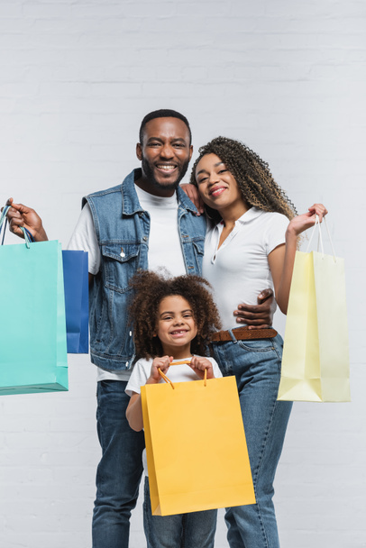 happy african american couple holding multicolored shopping bags while looking at camera on grey - Photo, Image