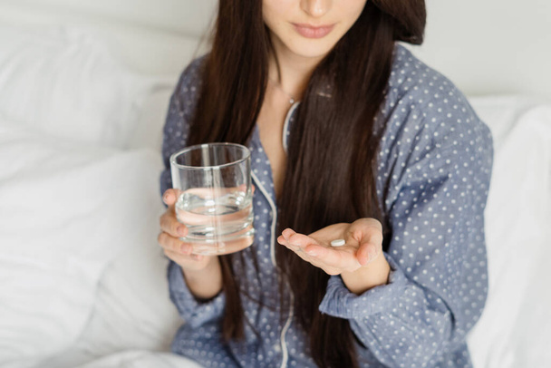 Cropped shot of brunette woman in pajama holding medication and glass of water while sitting on bed at home. Young lady having daily dose of health support according to prescription of doctor - 写真・画像