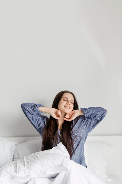 Guten Morgen. Glückliche junge Frau im Pyjama sitzt im bequemen Bett mit weißer Bettwäsche, nachdem sie am Wochenende aufgewacht ist, lächelt, während sie ihren Körper streckt und freut sich darauf, ihren Tag zu genießen, vertikale Aufnahme - Foto, Bild
