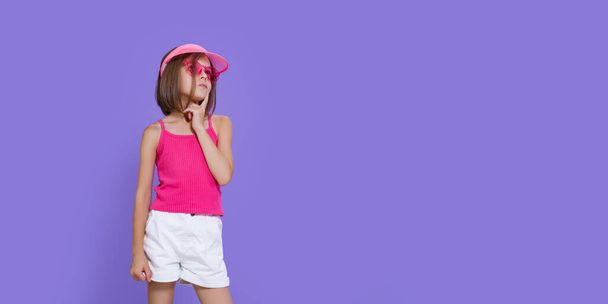 Little thoughtful girl in white shorts, a pink T-shirt, pink trendy glasses and a summer visor on a purple background in the studio - Foto, Imagem