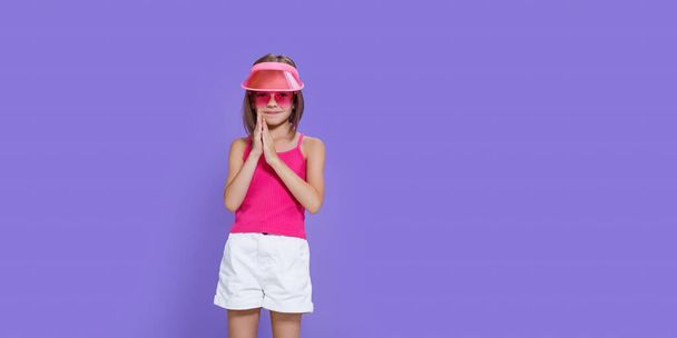 a little girl in white shorts, a pink T-shirt, pink trendy glasses and a summer visor on a purple background in the studio - Zdjęcie, obraz