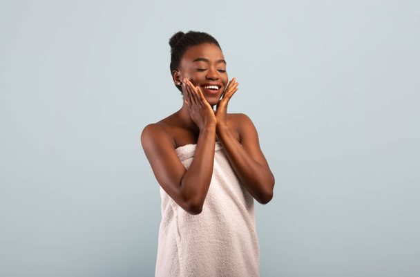Natürliches Konzept. Attraktive afrikanisch-amerikanische Dame berührt ihre weiche makellose Haut auf den Wangen, grauer Hintergrund - Foto, Bild