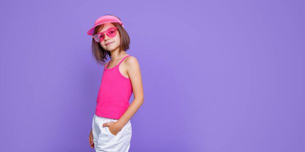 a little girl in white shorts, a pink T-shirt, pink trendy glasses and a summer visor on a purple background in the studio - Fotografie, Obrázek