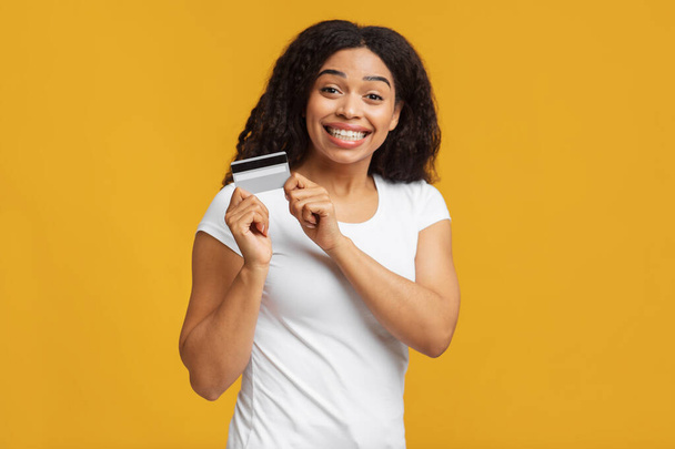 Ώρα για ψώνια. Πορτρέτο της αφρικανικής Αμερικής γυναίκα κρατώντας πιστωτική κάρτα και χαμογελώντας στην κάμερα, κίτρινο φόντο - Φωτογραφία, εικόνα