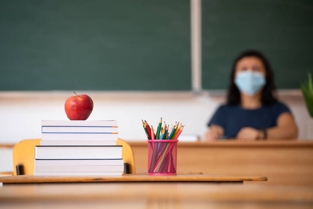 Back to School Concept. Apple, accessories and books in the classroom, teacher wearing mask in the background. - Photo, Image