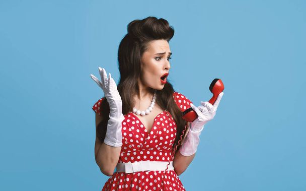 Σοκαρισμένος νεαρή γυναίκα pinup σε ρετρό φόρεμα κρατώντας σταθερό τηλέφωνο δέκτη, αίσθημα ενόχλησης για τηλεφωνική συνομιλία - Φωτογραφία, εικόνα