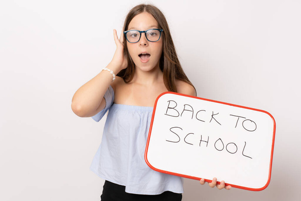 Lenyűgöző fiatal lány szemüveget visel, fehér táblát tart a fehér háttér felett. Vissza az iskolába. - Fotó, kép