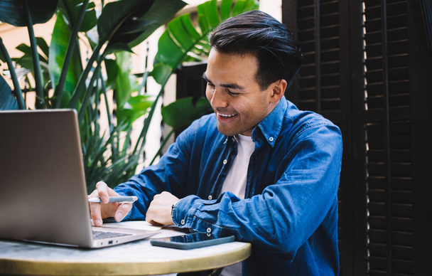 Joven freelancer masculino étnico sonriente con bolígrafo que navega por Internet en netbook mientras está sentado en la mesa en la cafetería de la ciudad en el día de verano - Foto, Imagen