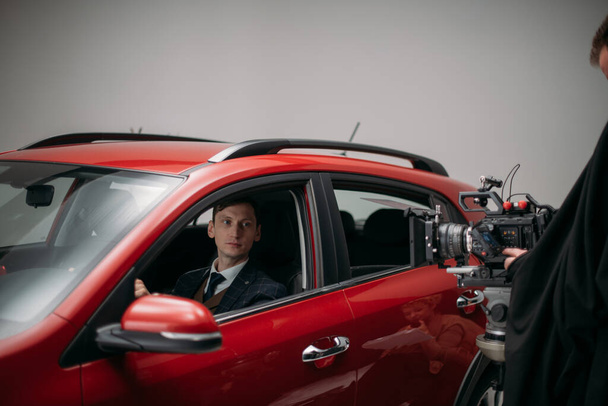 Professional male actor works in the frame on the set. Shooting with a car on a large white cyclorama. Handsome young man on the set of a movie, commercial or TV series. Filming indoors, studio - Photo, Image