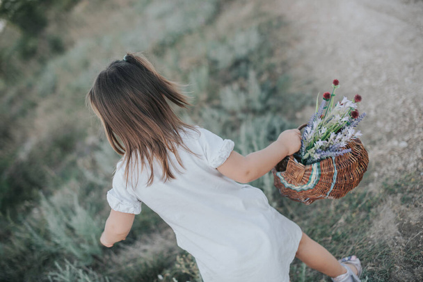 A little girl in a white dress with a wicker basket with wildflowers. - Фото, изображение