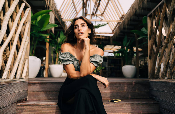 Pensive tender female in trendy outfit leaning chin on hand while relaxing on steps leading to wooden terrace of cozy cafe with plant decoration in summer day - Foto, Imagen