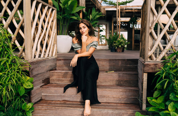 Calm tender barefoot woman in stylish clothes leaning head on hand resting alone on exterior steps of wooden terrace decorating green plants against cozy interior of summer cafe in daylight - Foto, Bild