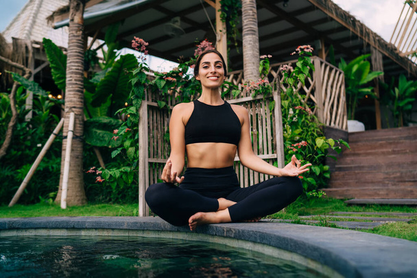 Portrait of happy Caucasian woman in sportswear posing in lotus pose during meditation time at pool terrace, cheerful female yogi smiling at camera feeling carefree while practice zen harmony - Fotó, kép