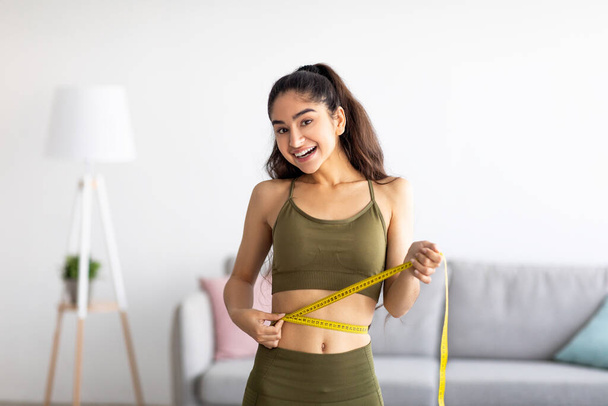 Successful weight loss. Slim Indian woman measuring waist with tape, satisfied with slimming program at home - 写真・画像