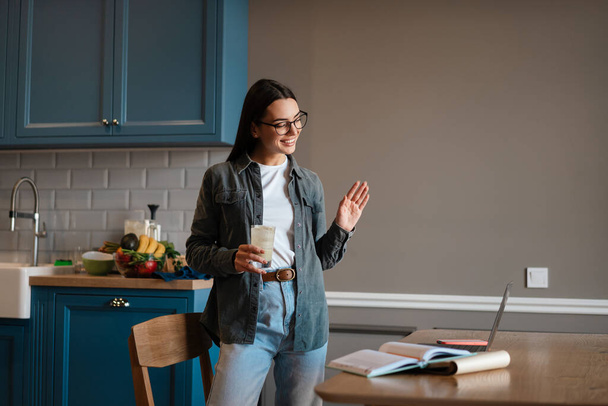 Happy mid aged brunette woman holding glass with beverage while standing at the kitchen table, on a video call via laptop computer, waving hand - 写真・画像