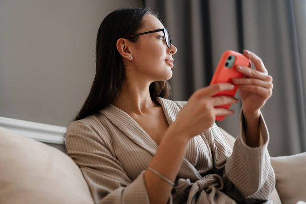 White young woman using mobile phone while sitting on couch at home - Foto, Bild