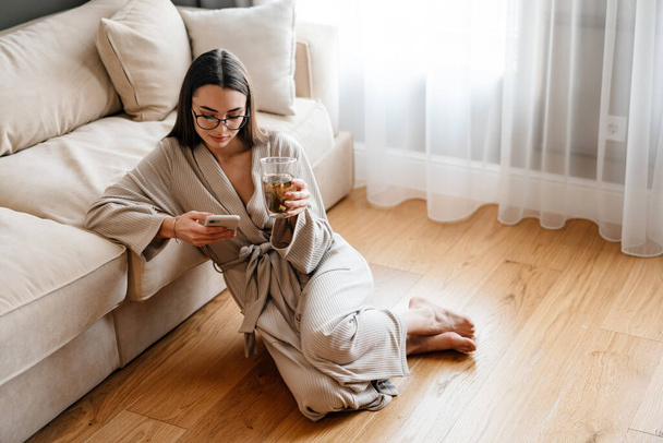 White woman drinking tea and using mobile phone while sitting on floor at home - Фото, зображення