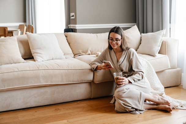 White woman drinking tea and using mobile phone while sitting on floor at home - Fotoğraf, Görsel