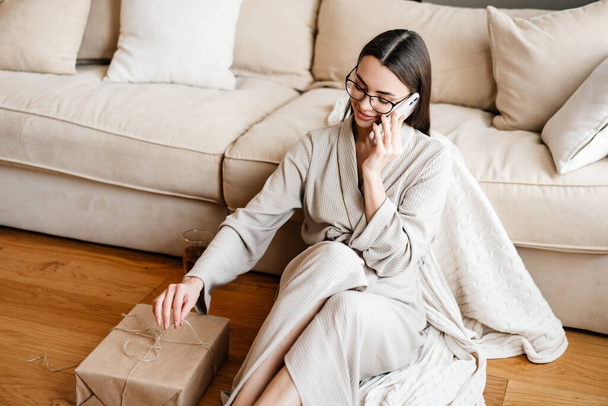 White woman talking on mobile phone while sitting with gift box at home - Photo, Image
