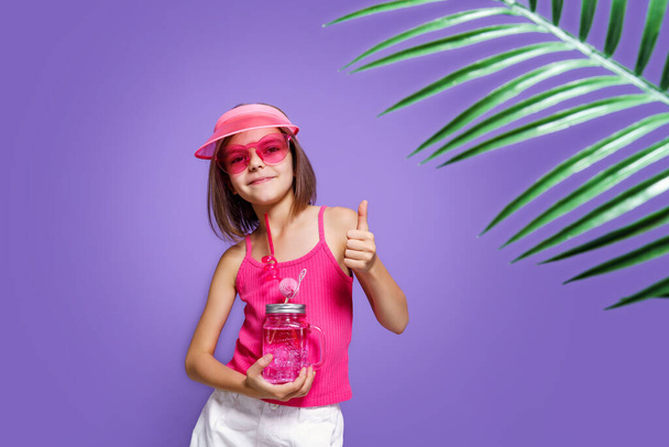 a little girl in white shorts, a pink T-shirt, pink trendy glasses, with a drink in hand and a summer visor on a purple background in the studio - Photo, Image