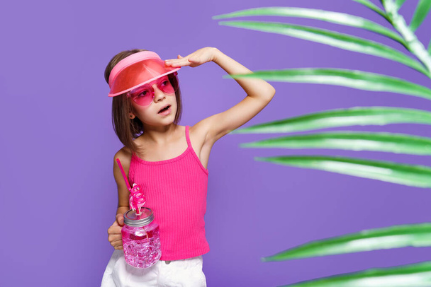 a little girl in white shorts, a pink T-shirt, pink trendy glasses, with a drink in hand and a summer visor on a purple background in the studio - Φωτογραφία, εικόνα