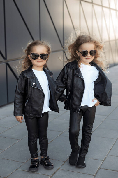 Two sisters in same outfits: white t-shirts, leather black jackets and leggings, sunglasses hearts. - Фото, изображение