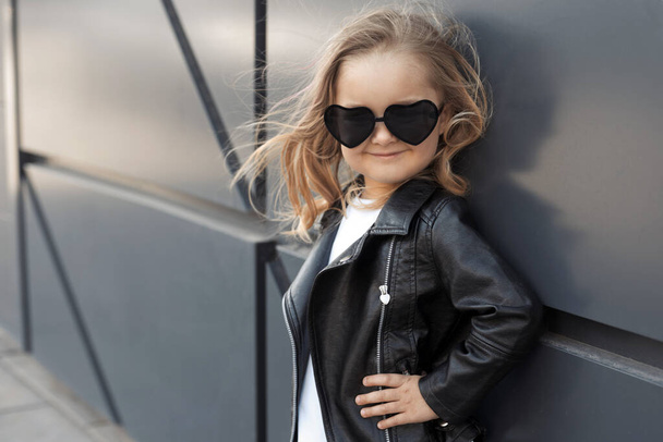 Pretty baby girl in fashion outfit: white t-shirt, leather black jacket and leggings, sunglasses hearts - Foto, Imagen