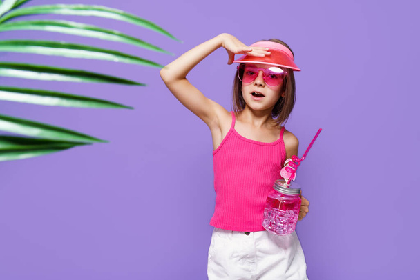 a little girl in white shorts, a pink T-shirt, pink trendy glasses, with a drink in hand and a summer visor on a purple background in the studio - Fotografie, Obrázek